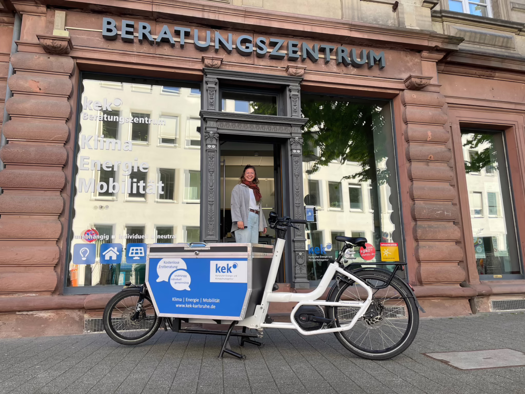 Foto vom Beratunhszentrum der KEK (denkmalgeschütztes Gebäude) mit einem weißen Lastenrad davor, auf dem groß das Logo der KEK zu sehen ist. Im EIngang steht eine Perosn und lächelt.