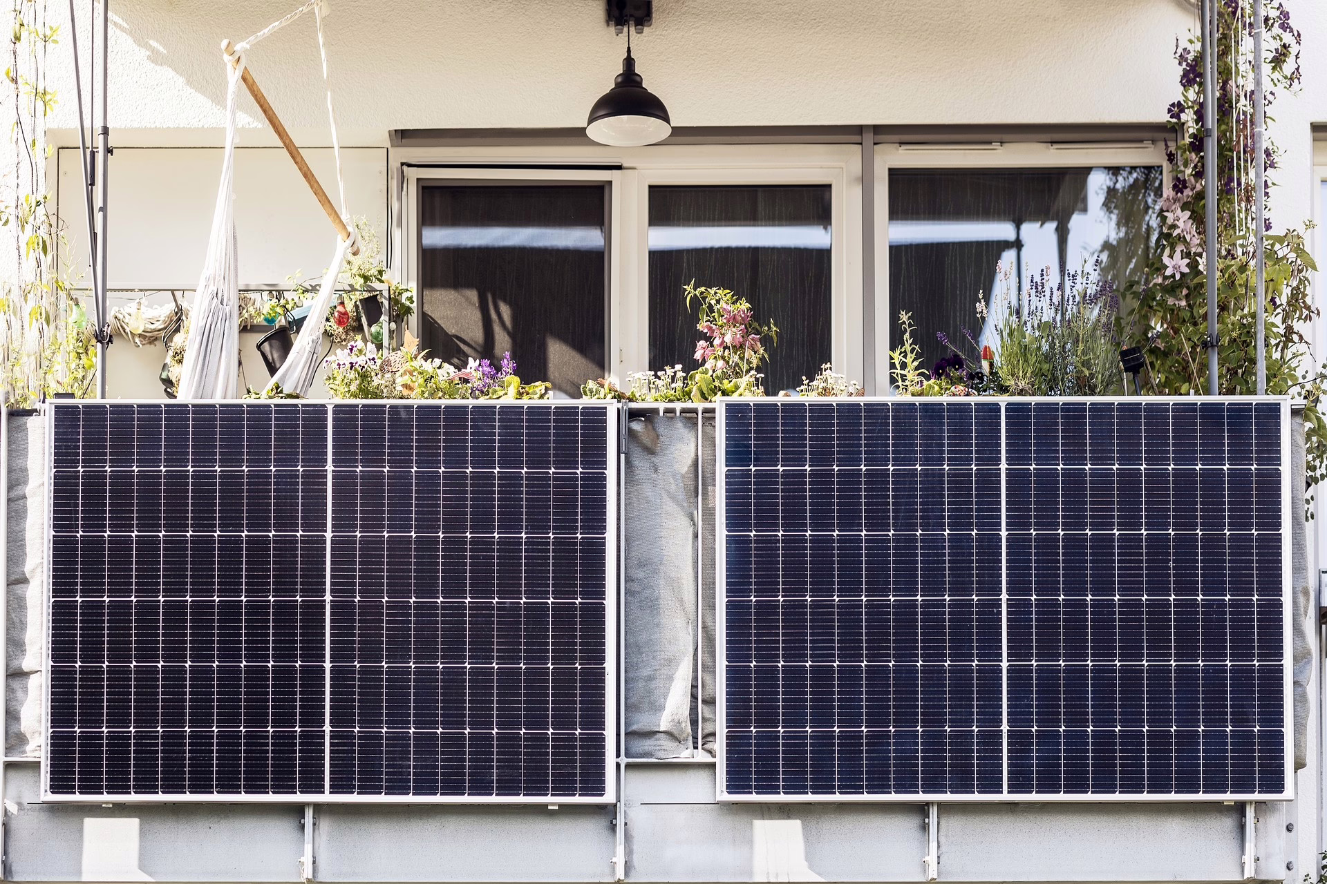Frontale Draufsicht auf ein Balkongeländer, an dem zwei parallel befestigte Balkon-Photovoltaik-Panele befestigt sind. Auf dem Balkon befinden sich mehrere Pflanzen an allen Seiten sowie eine an der Decke befestigte Sitzgelegenheit.