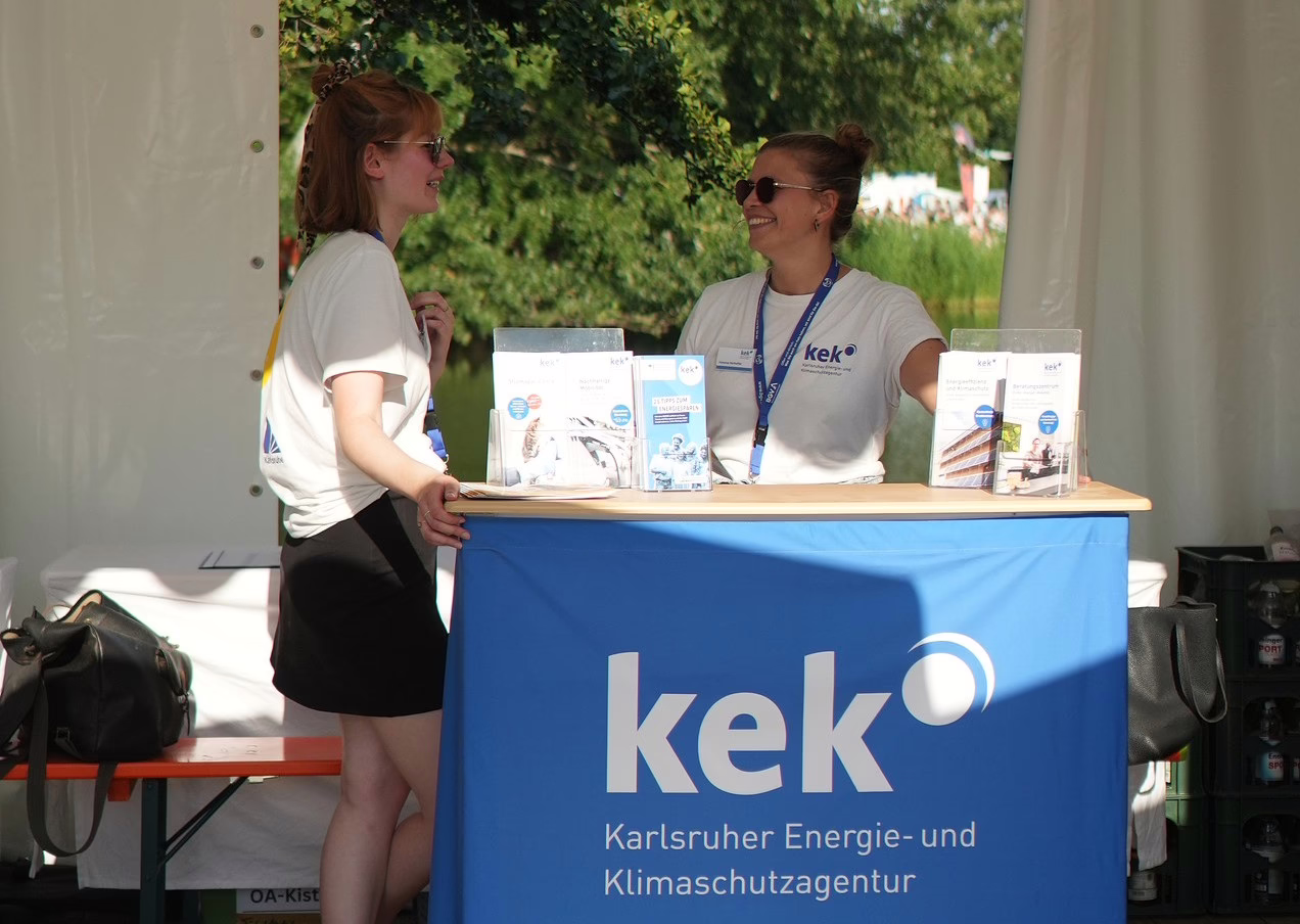 Zwei Frauen stehen an einem Infostand der KEK. Vorne ist eine Theke mit dem KEK-Logo.