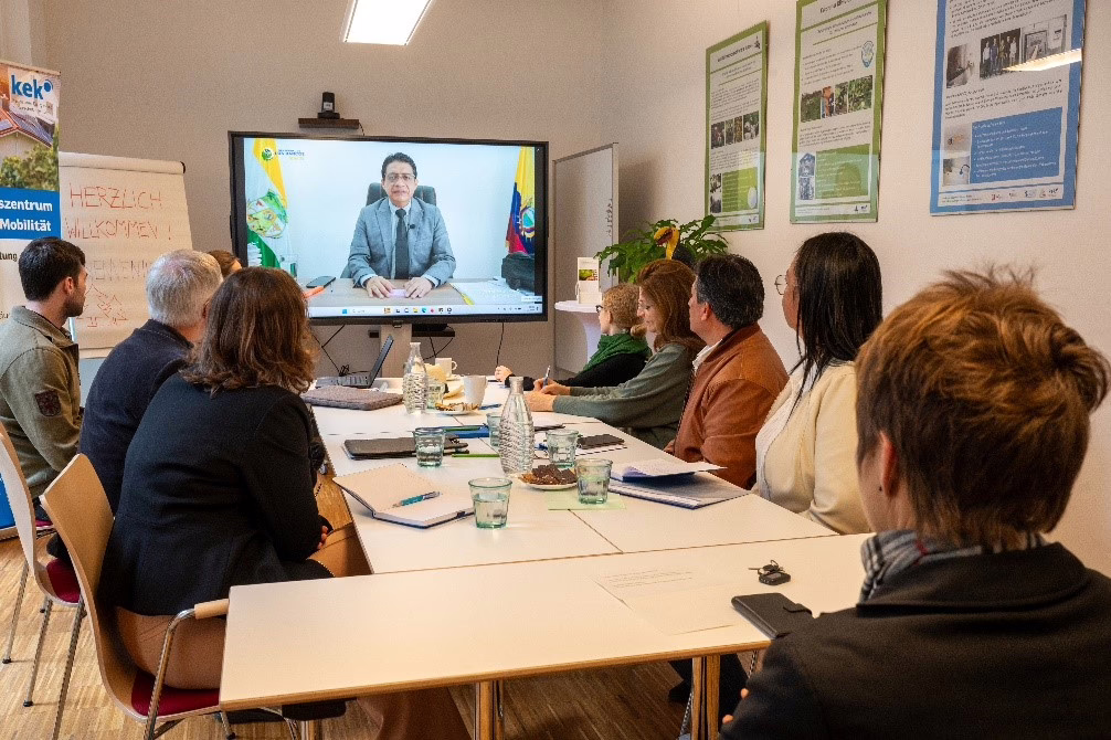 Eine Gruppe Menschen sitzt an einem großen Tisch zusammen, auf einem Bildschirm ist jemand per Video zugeschaltet. Es war der Delegationsbesuch aus Ecuador.