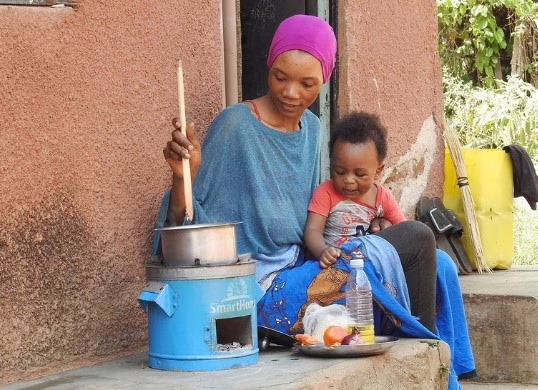 Das Bild wurde in Uganda aufgenommen. Eine Mutter sitzt mit ihrem Kind vor einer Lehmhütte. Mit einem kleinen Kochofen bereitet sie Essen zu.