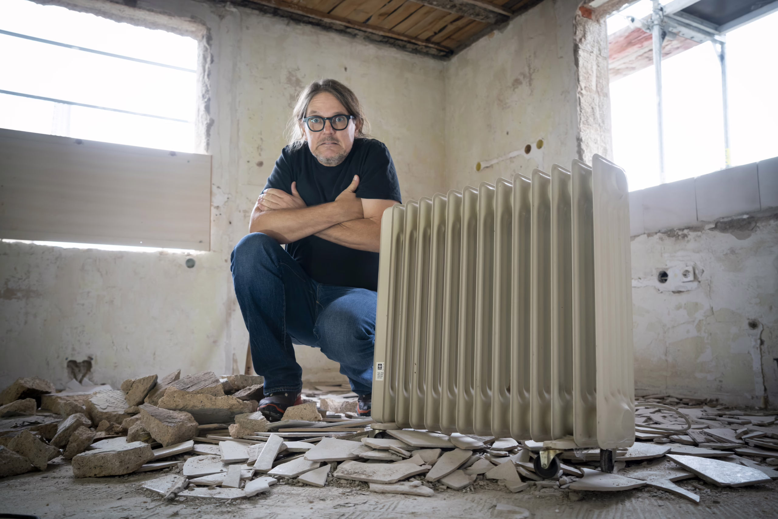 Foto vom Energiesparkommissar (Carsten Herbert), der in einem kapputten Haus neben einer Heizung sitzt und die Arme verschränkt.