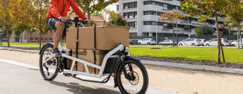 Männliche Person sitzt auf einem Lastenrad, auf dem zwei große Pakete befestigt sind. Im Hintergrund ist