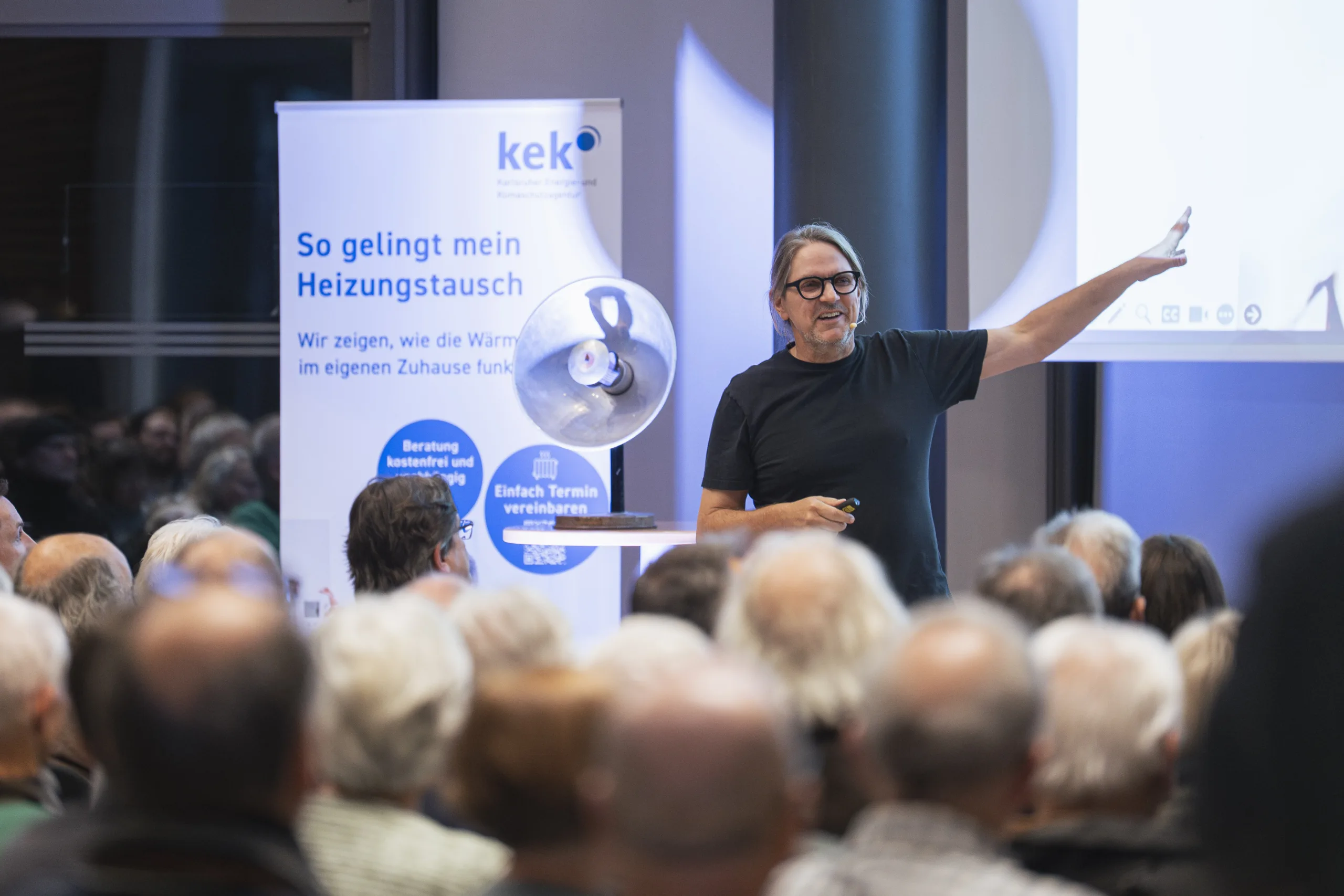 Energiesparkommissar Carsten Herbert zeigt auf seine PowerPoint Präsentation. Im hintergrund das KEK Roll-up und eine Lampe.