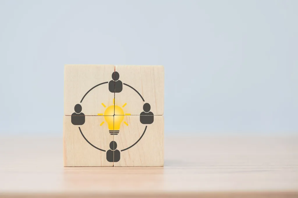 Vier Holzklötze, die in einem Quadrat gestapelt sind. In der Mitte ist eine Glühbirne, drumherum vier Personen angeordnet, die durch einen Kreis miteinander verbunden sind.