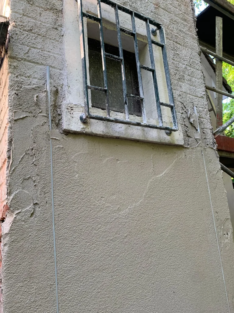 Ein Fenster Außen ist zu sehen. An der unteren Seite der Fassade wurde der Kalkdämmputz bereits angebracht, im oberen Teil noch nicht. 