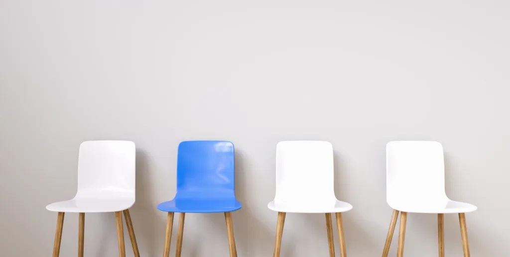 Vier Stühle stehen nebeneinander. Drei sind weiß, einer ist blau.