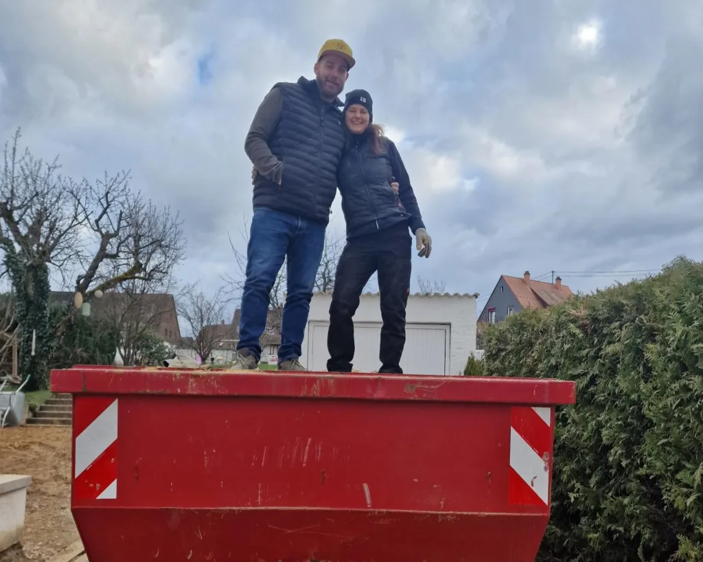 Sarah und ihr Mann stehen in Arbeitsklamotten in einem roten Container