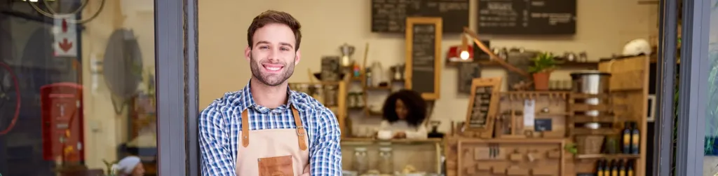 Mann mit brauner Schürze, der im Türrahmen eines Ladengeschäfts steht.
