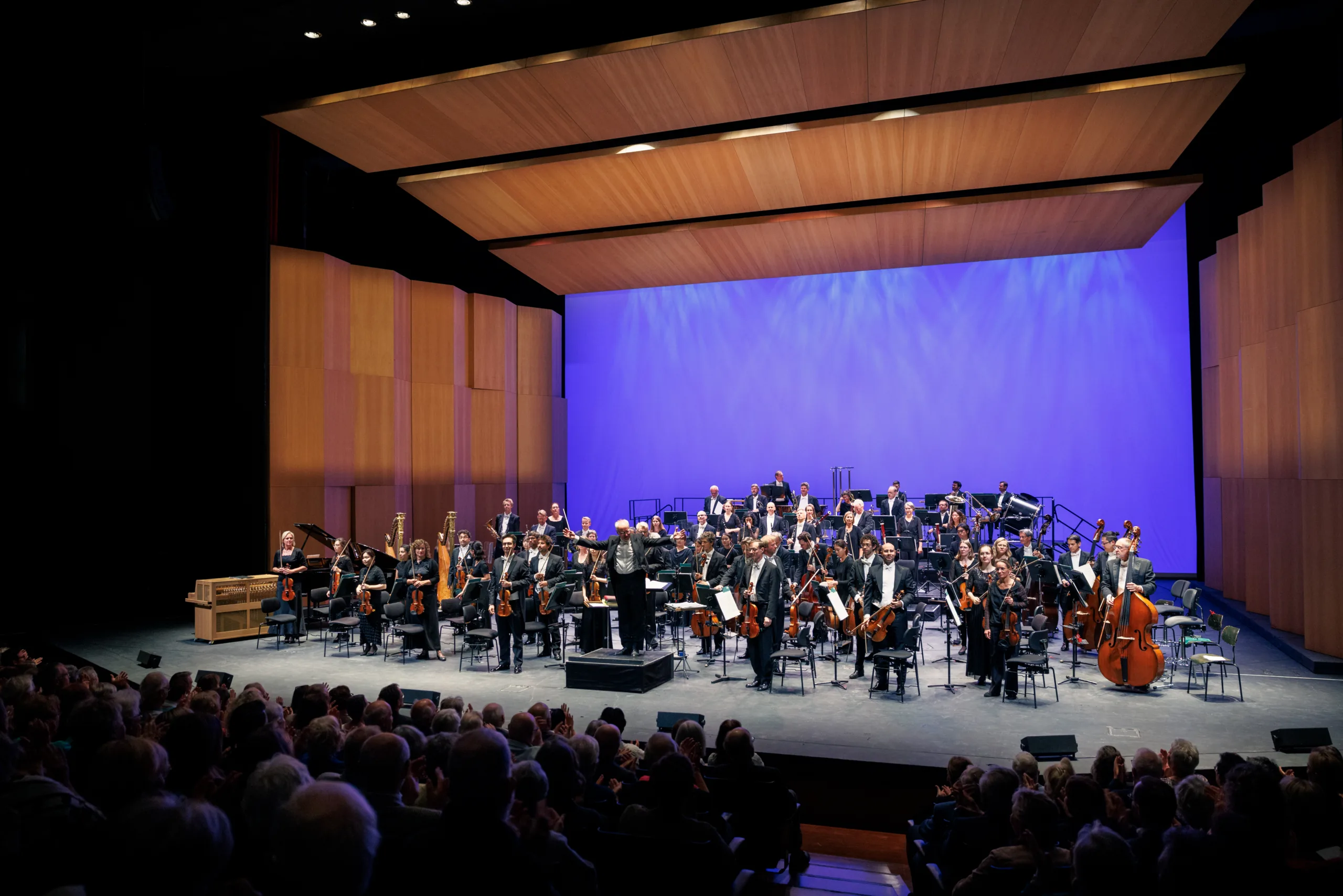 Die badische Staatskapelle ist auf einer Bühne zu sehen. Der Dirigent und die Musikerinnen und Musiker stehen am Ende des Konzerts und schauen lächelnd ins Publikum