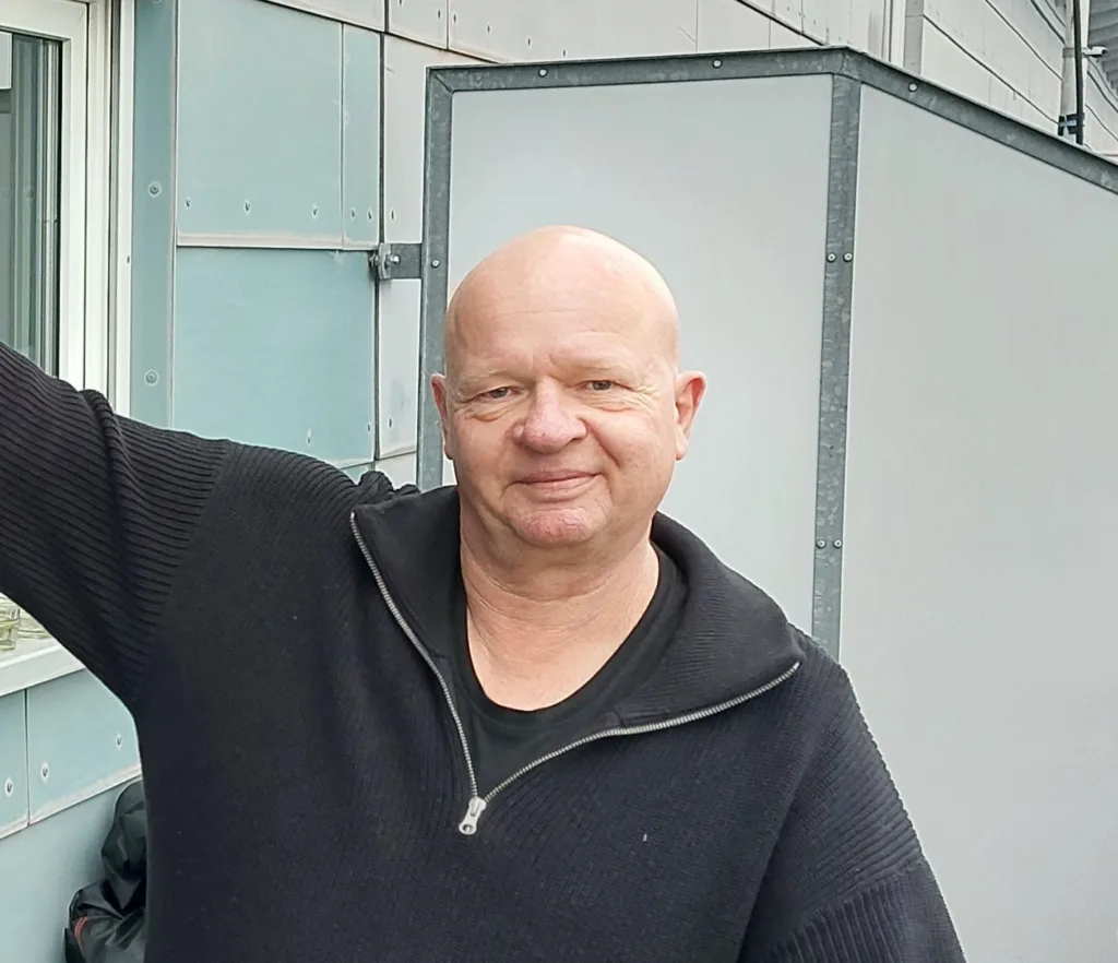 Stefan lächelt in die Kamera. Er steht auf seinem Balkon. 