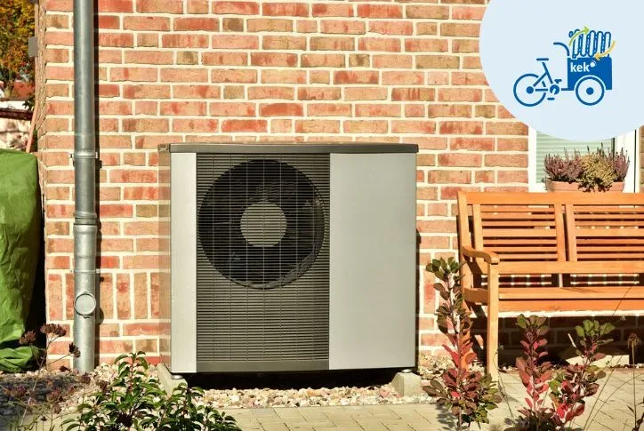 Foto eines Backsteinhauses, vor dem eine graue Wärmepumpe steht. Rechts daneben steht eine braune Hokzbank.