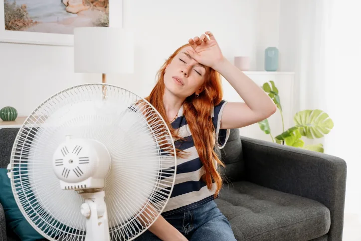 Frau sitzt vor einem Ventilator und hät ihre Hand an die Stirn.